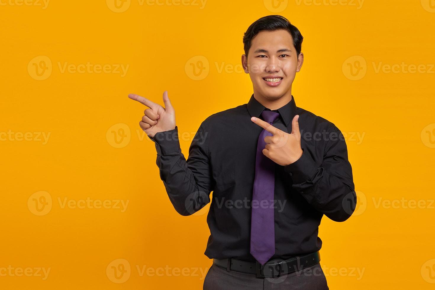 Smiling young Asian man pointing fingers at copy space on yellow background photo