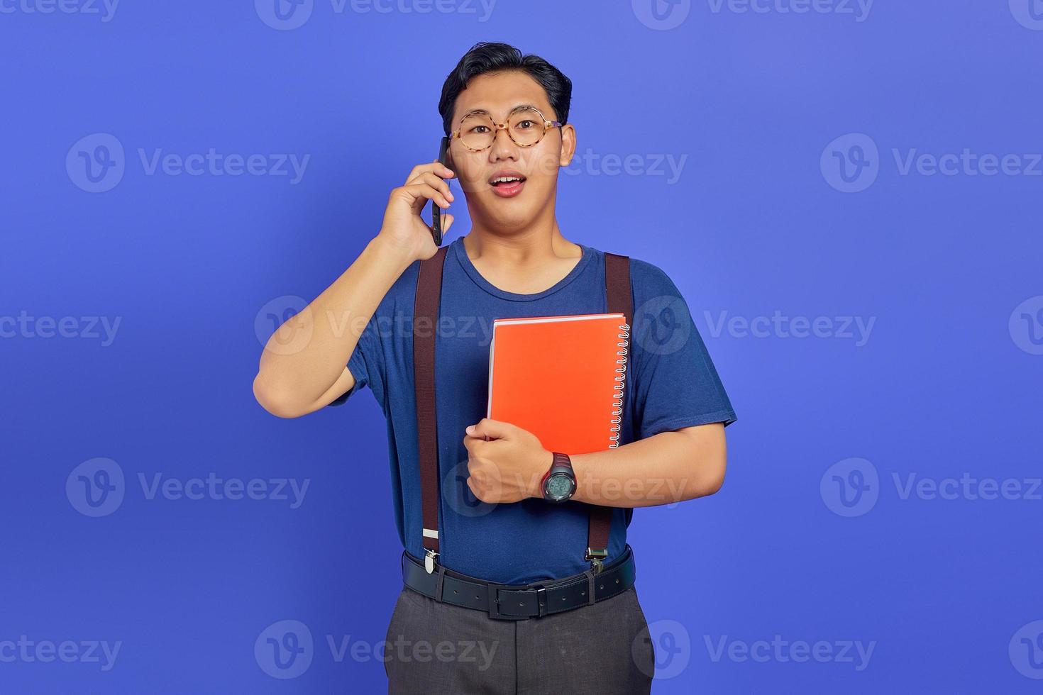 Joven estudiante sorprendido hablando por teléfono inteligente mientras sostiene el cuaderno sobre fondo púrpura foto