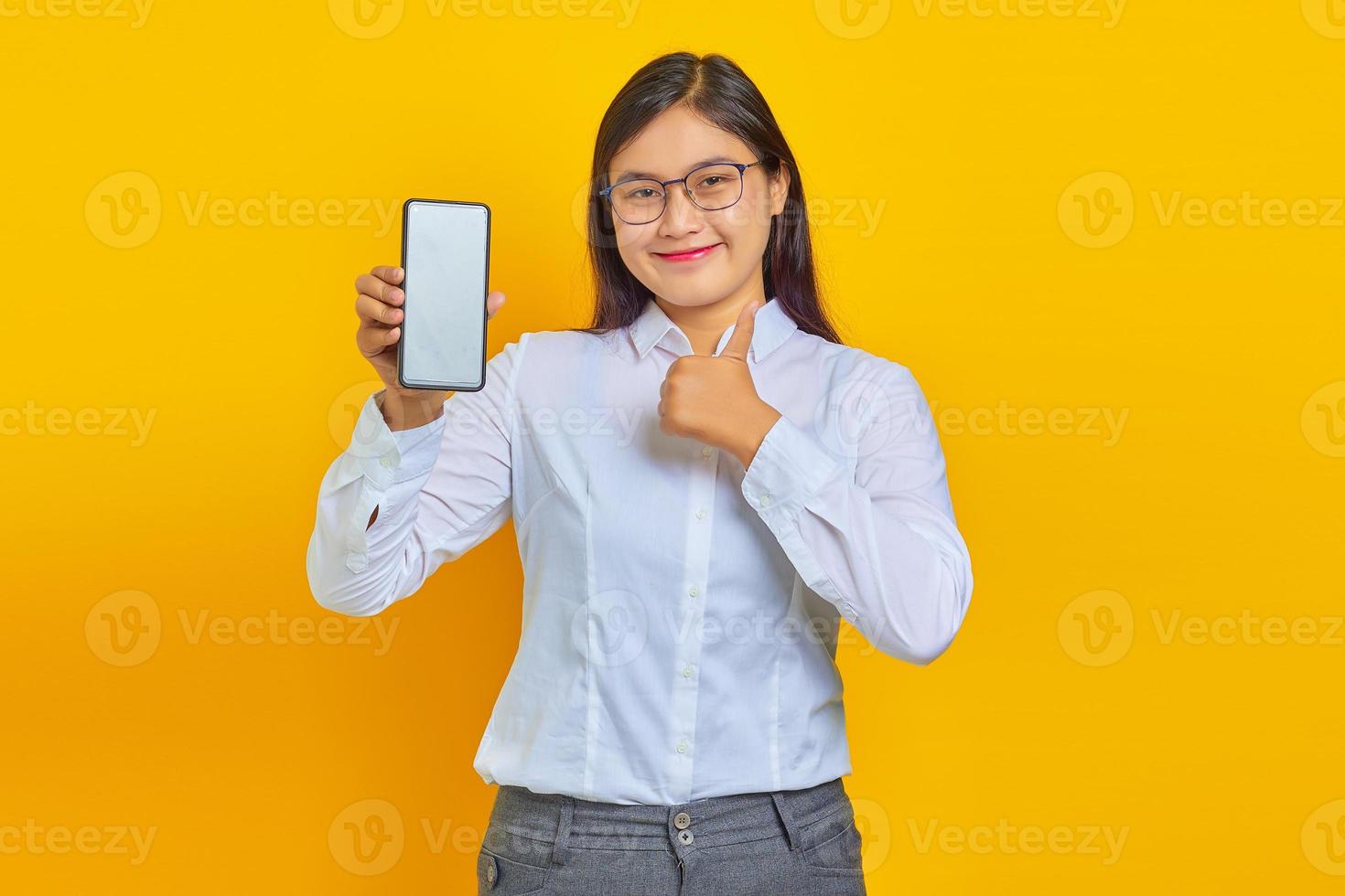 Mujer asiática emocionada y alegre que muestra la pantalla del teléfono inteligente en blanco y los pulgares hacia arriba sobre fondo amarillo foto