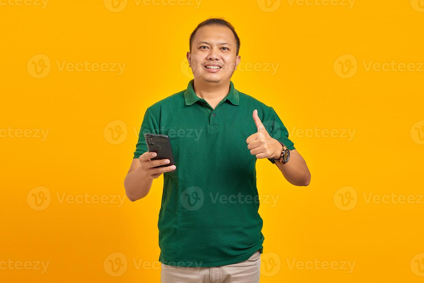 Portrait of cheerful Asian young man holding mobile phone and showing thumbs up sign over yellow background photo