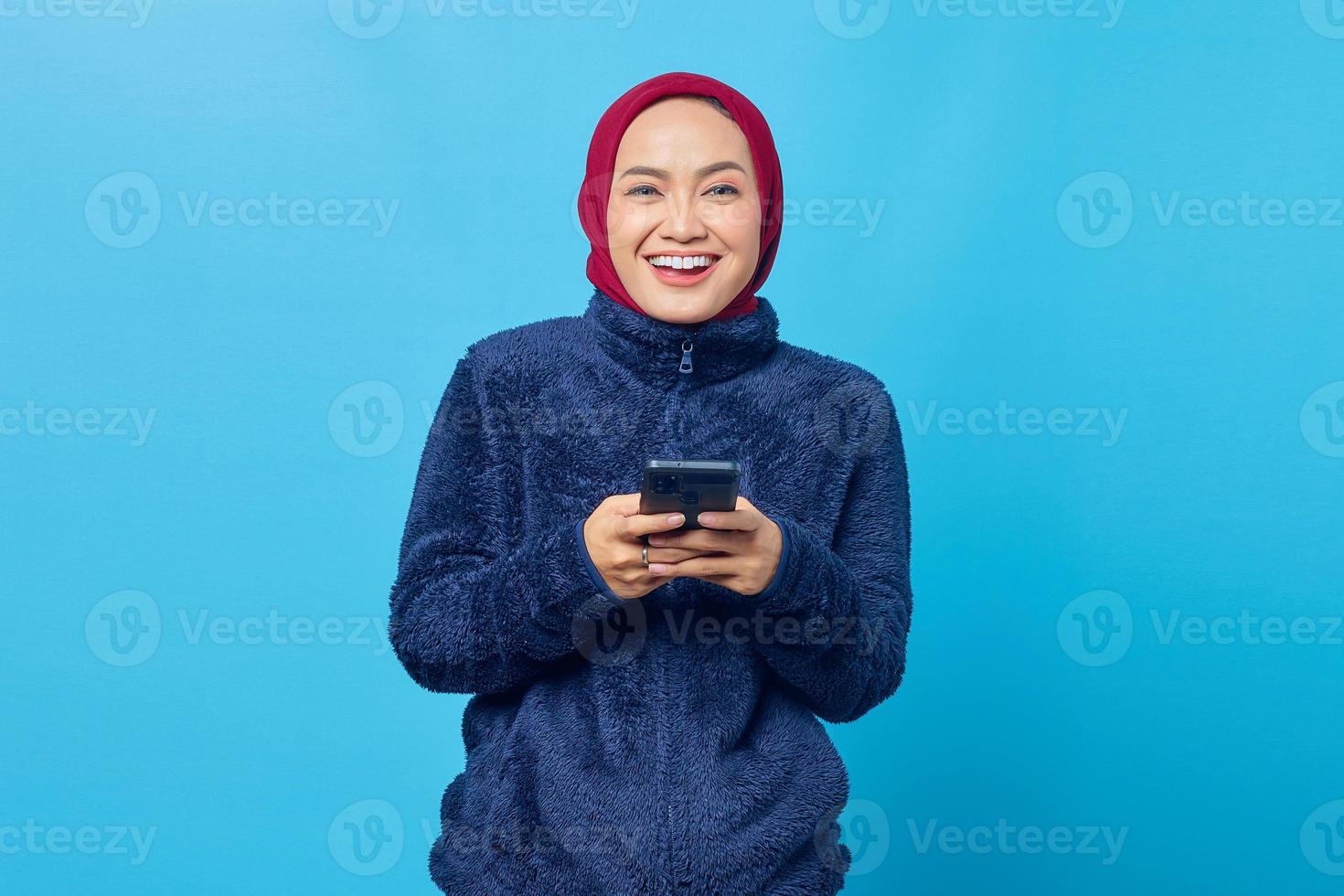 Cheerful young Asian woman using smartphone and looking at camera on blue background photo