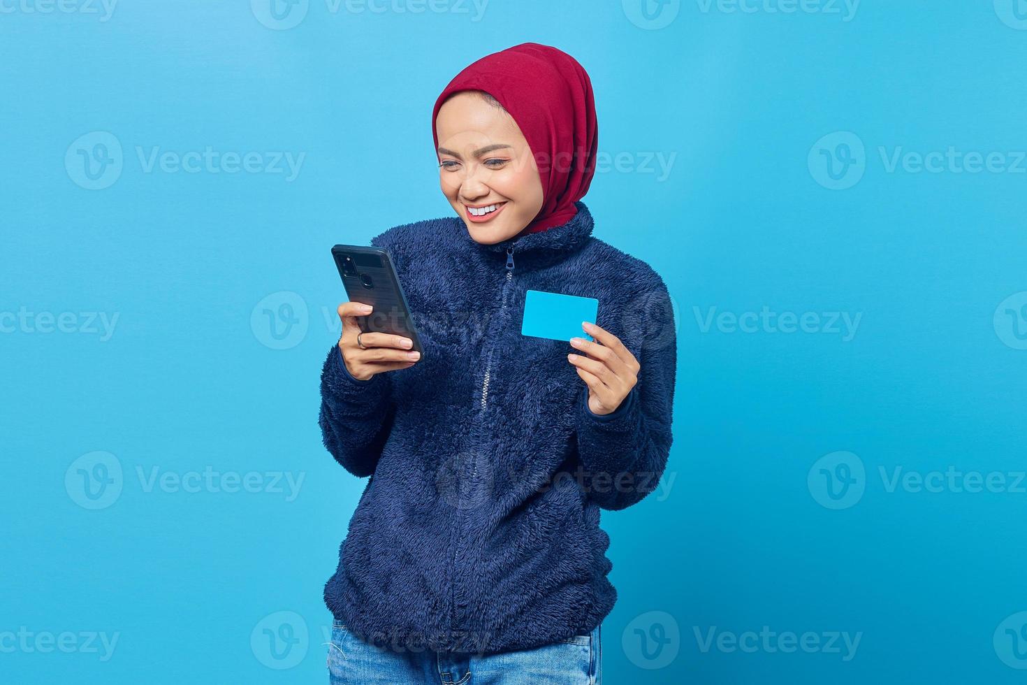 Alegre joven mujer asiática sosteniendo teléfono móvil y mostrando tarjeta de crédito sobre fondo azul. foto