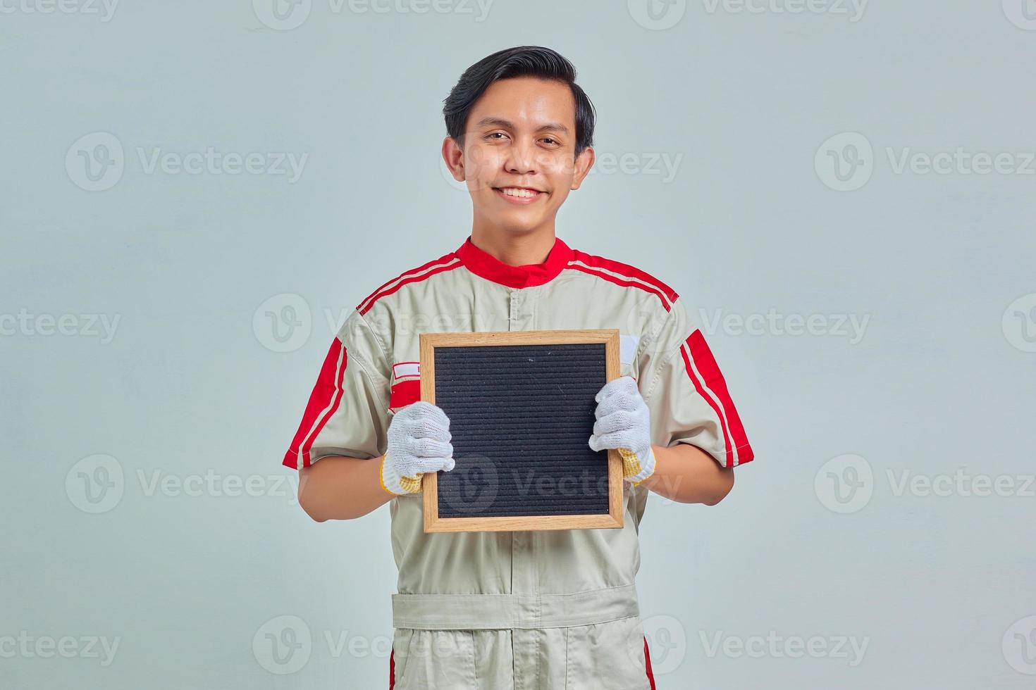 Portrait of cheerful young mechanic wearing uniform showing blank board on gray background photo