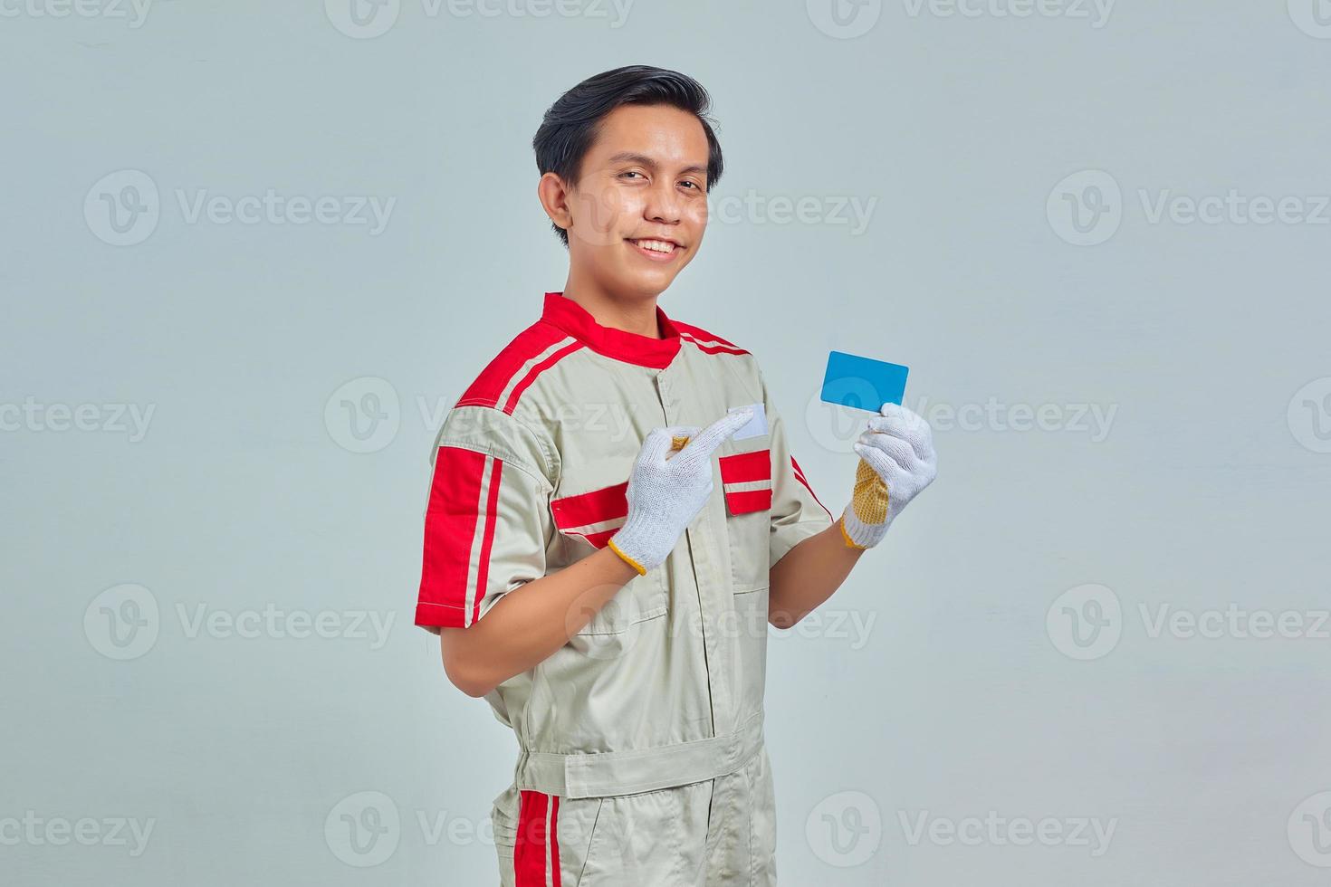 Apuesto joven mecánico apuntando a la tarjeta de crédito con el dedo con expresión sonriente sobre fondo gris foto