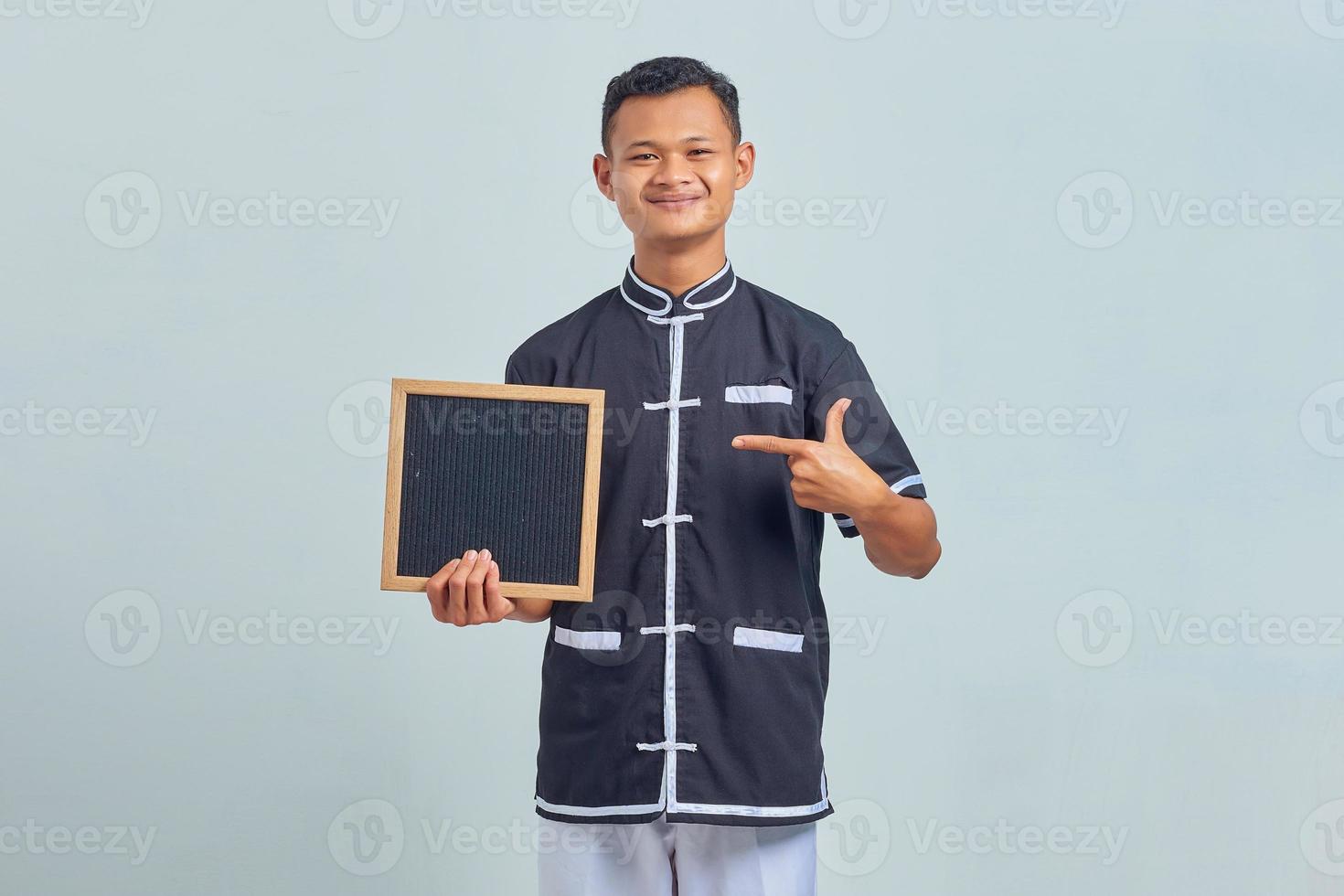 Portrait of cheerful asian young man wearing karate uniform pointing at blank board with finger on gray background photo