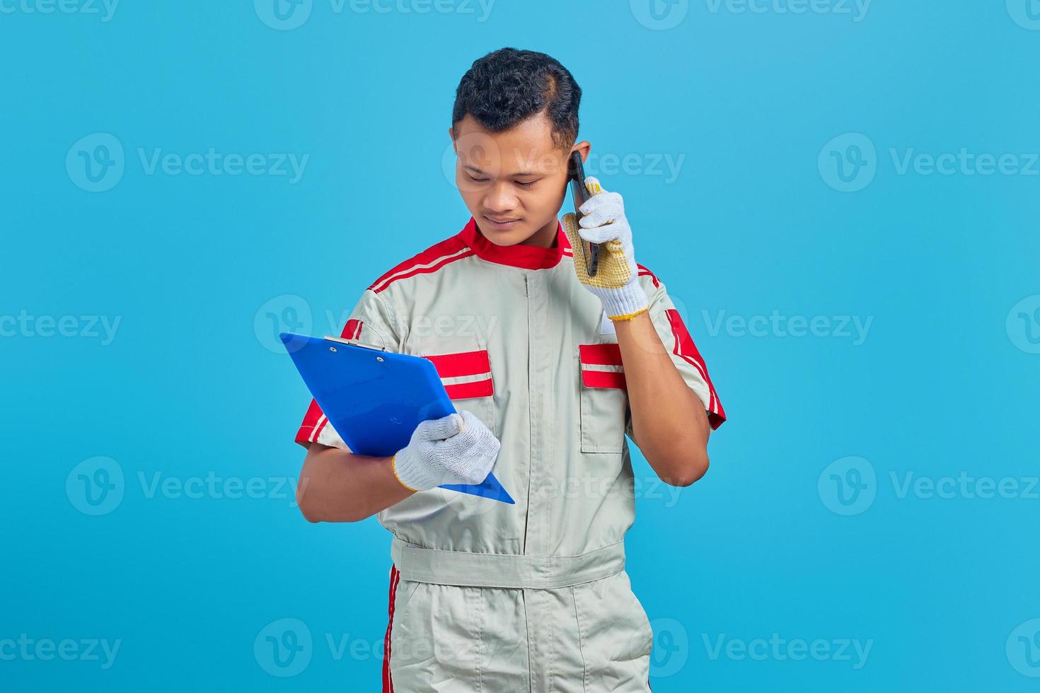 Serious looking young Asian mechanic talking on mobile and checking notes on clipboard isolated on blue background photo