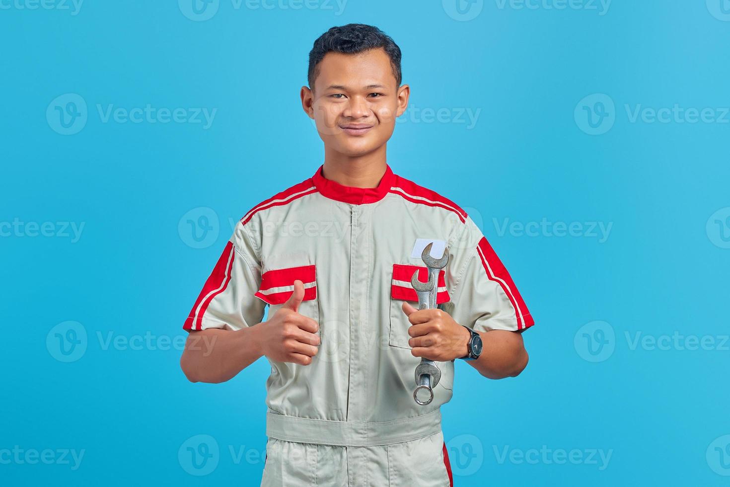 Portrait of smiling young Asian mechanic holding wrench and showing thumbs up to camera isolated on blue background photo