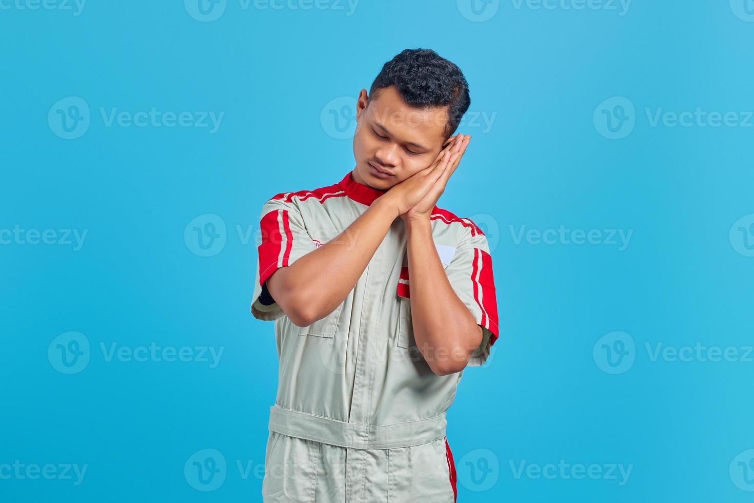 Portrait of cheerful handsome man wearing mechanical uniform with sleeping gesture on blue  background photo