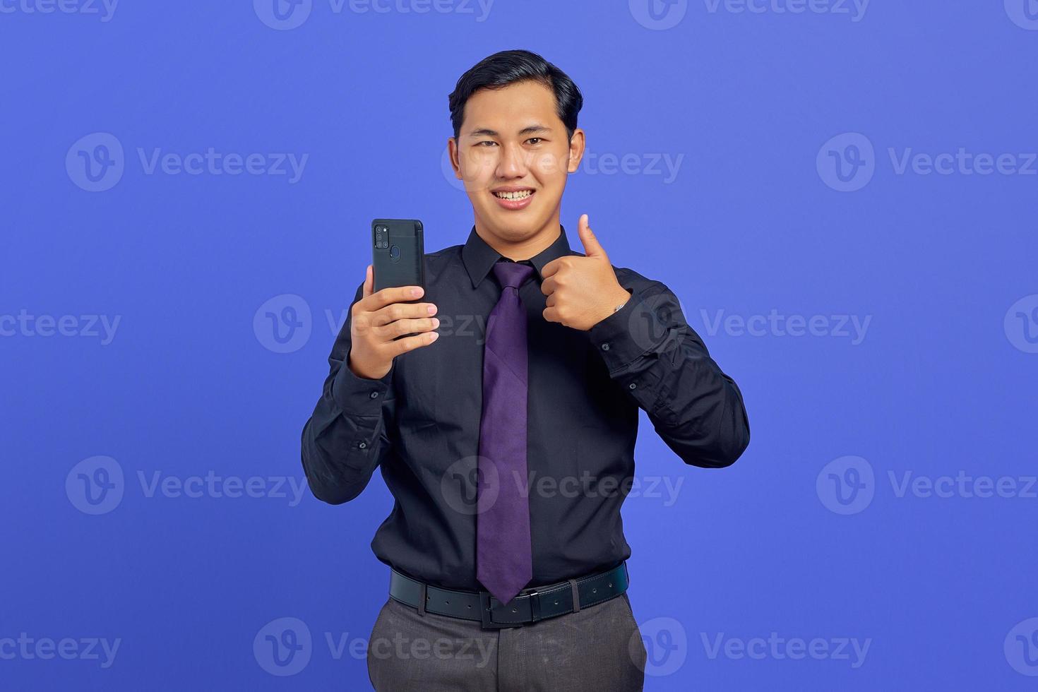 Happy handsome young businessman holding smartphone and giving thumbs up photo