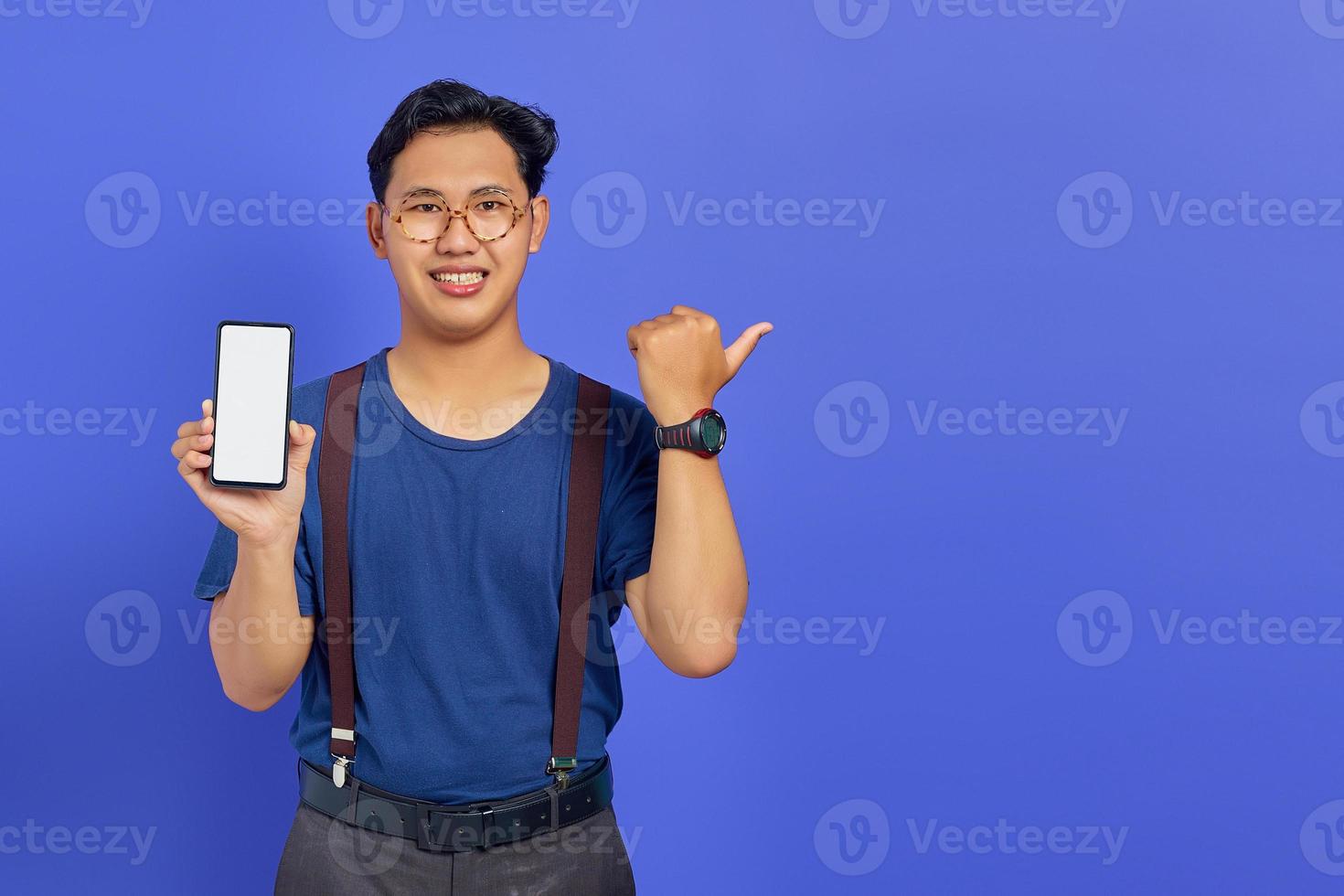 Cheerful asian young man pointing sideways in copy room and holding mobile phone isolated over purple background photo