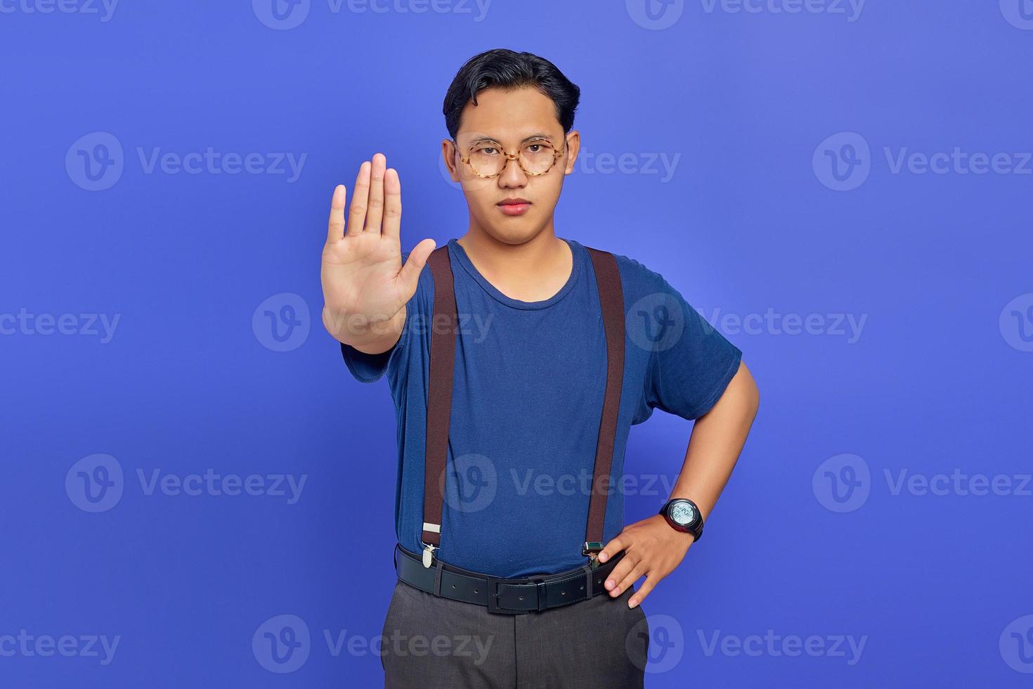 Portrait of handsome young man wearing blue shirt looking angry and doing stop gesture with palms on purple background photo