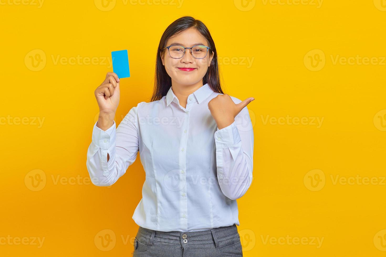 Smiling young Asian woman pointing finger at copy space and holding credit card on yellow background photo