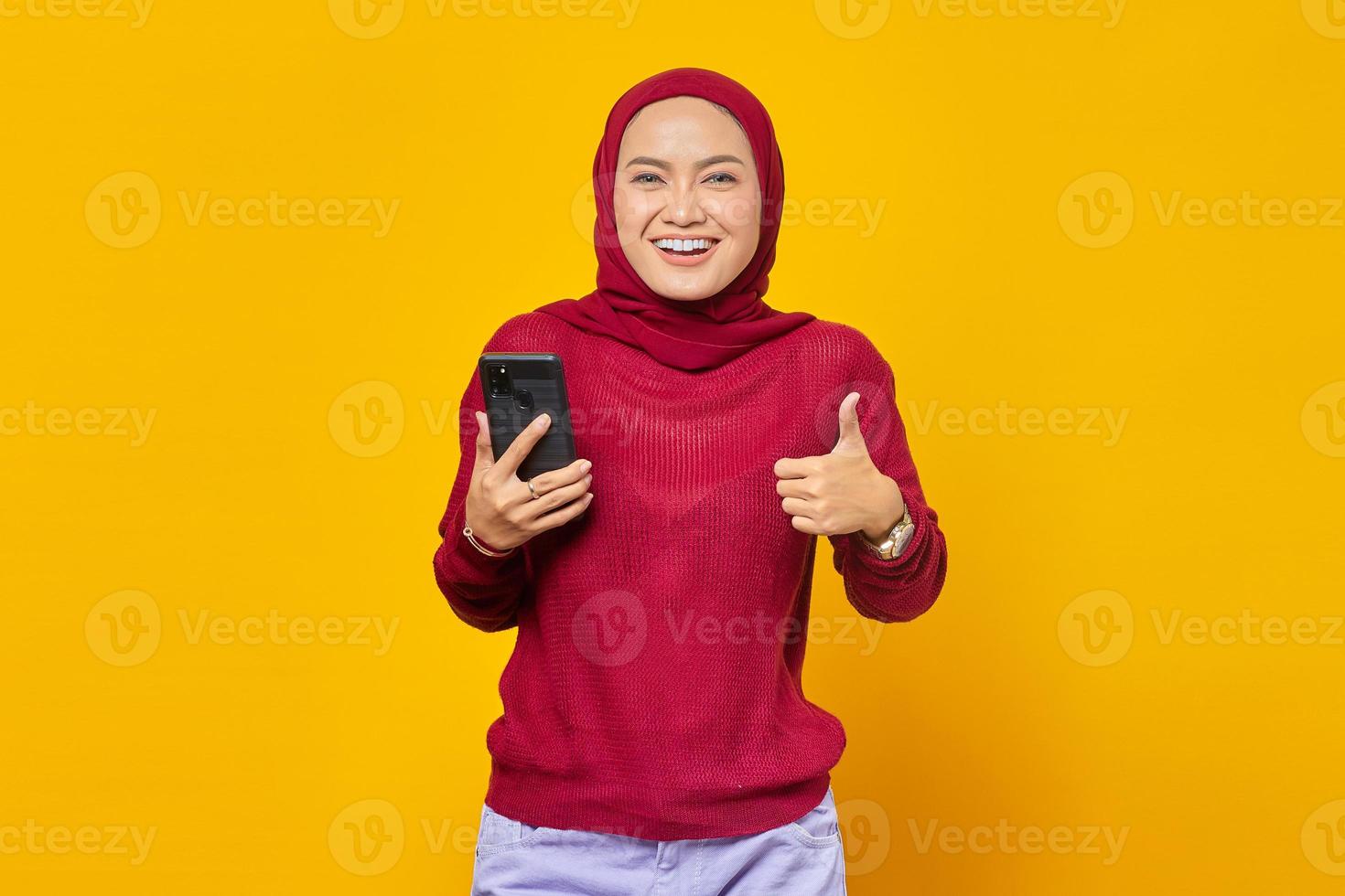 retrato, de, alegre, joven, mujer asiática, tenencia, teléfono móvil, y, actuación, pulgares arriba, señal, aislado, en, fondo amarillo foto