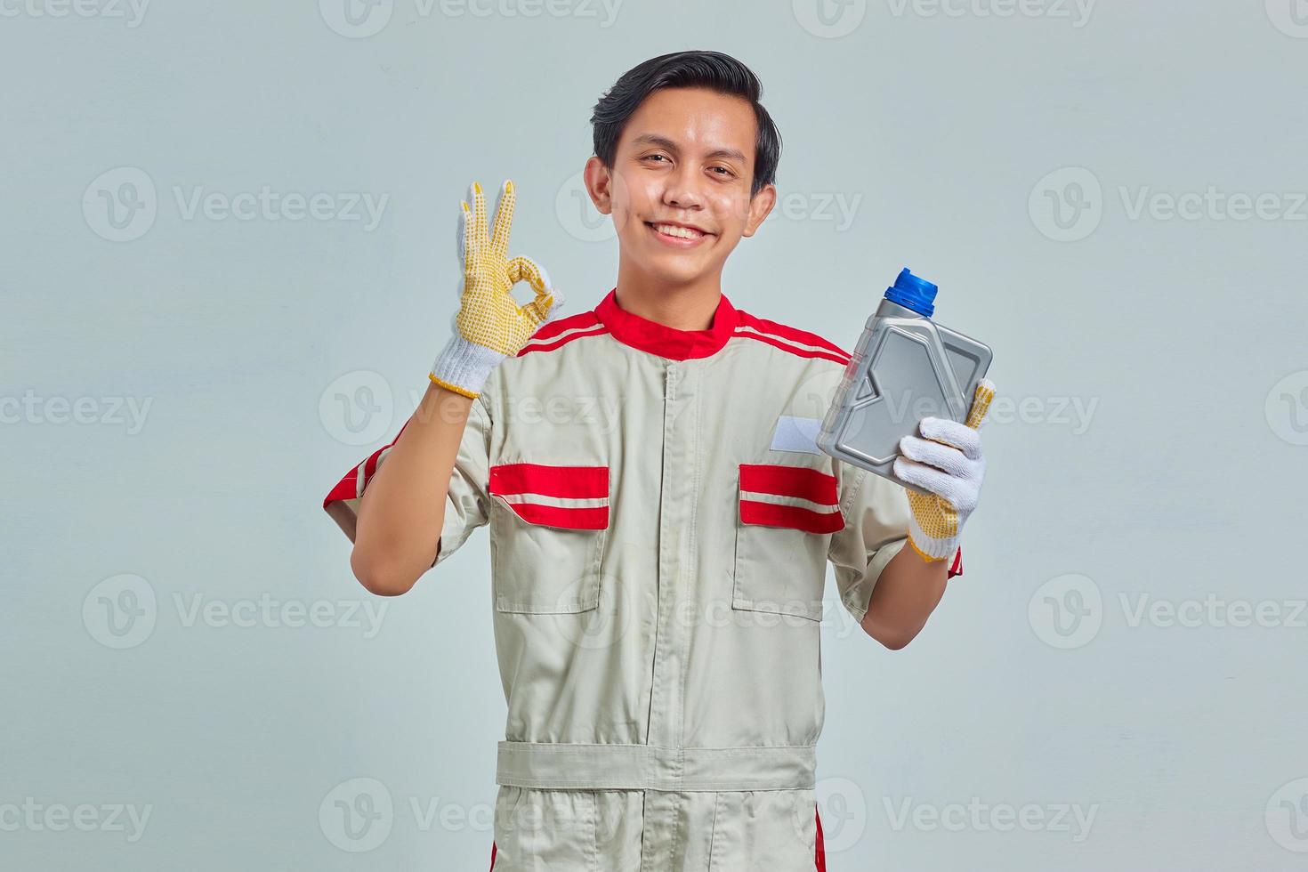 Retrato de smilling guapo vestido con uniforme de mecánico sosteniendo una botella de plástico de aceite de motor y mostrando aprobación con el pulgar hacia arriba sobre fondo gris foto