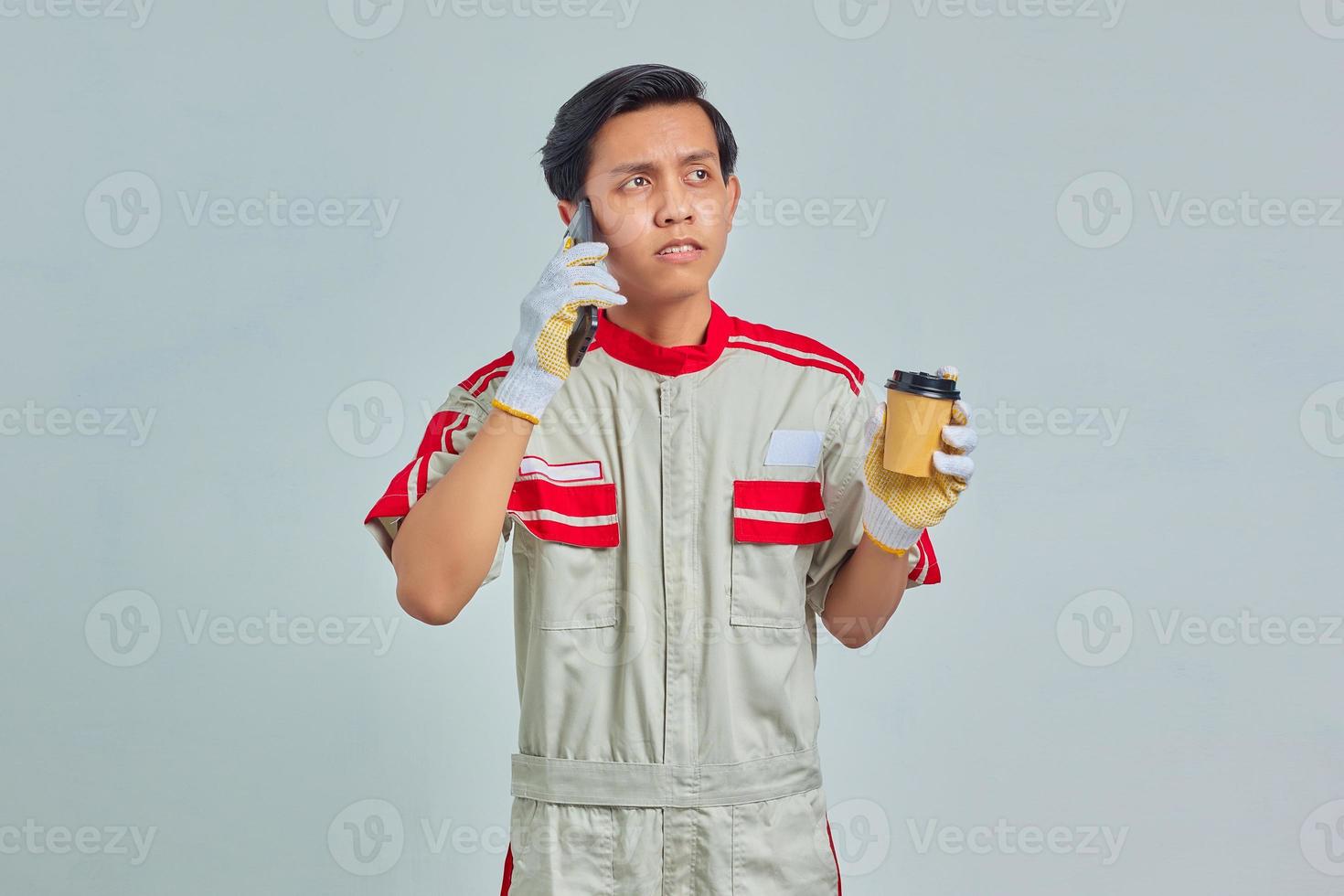 Surprised young mechanic talking on smartphone and holding coffee on gray background photo