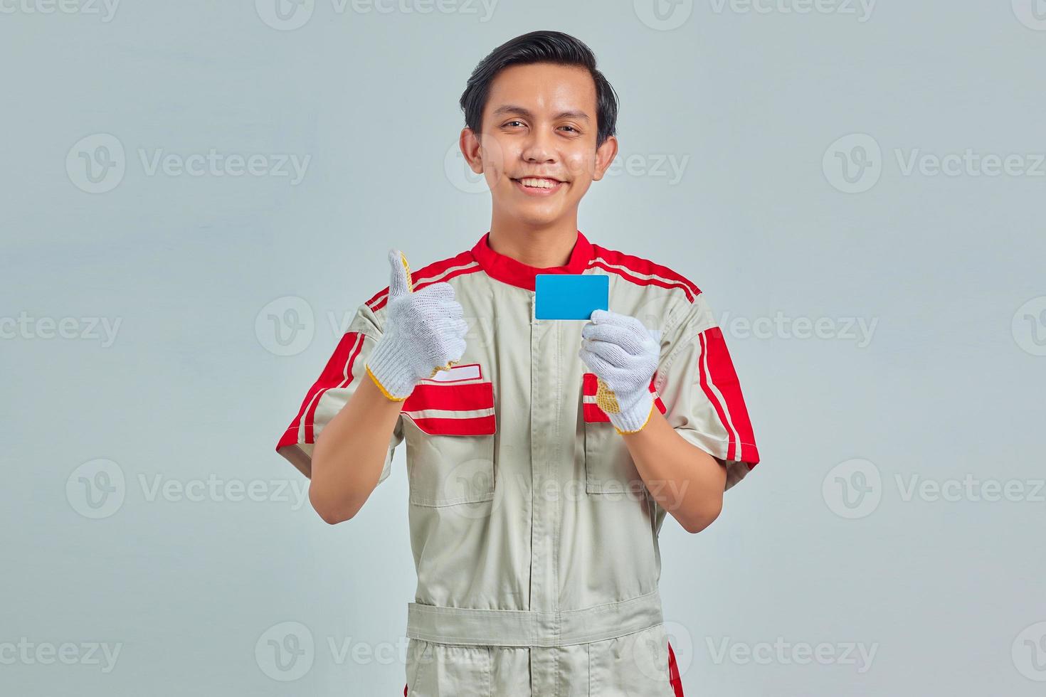 retrato, de, guapo, joven, mecánico, actuación, tarjeta de crédito, en, mano, y, pulgar arriba, gesto, en, fondo gris foto