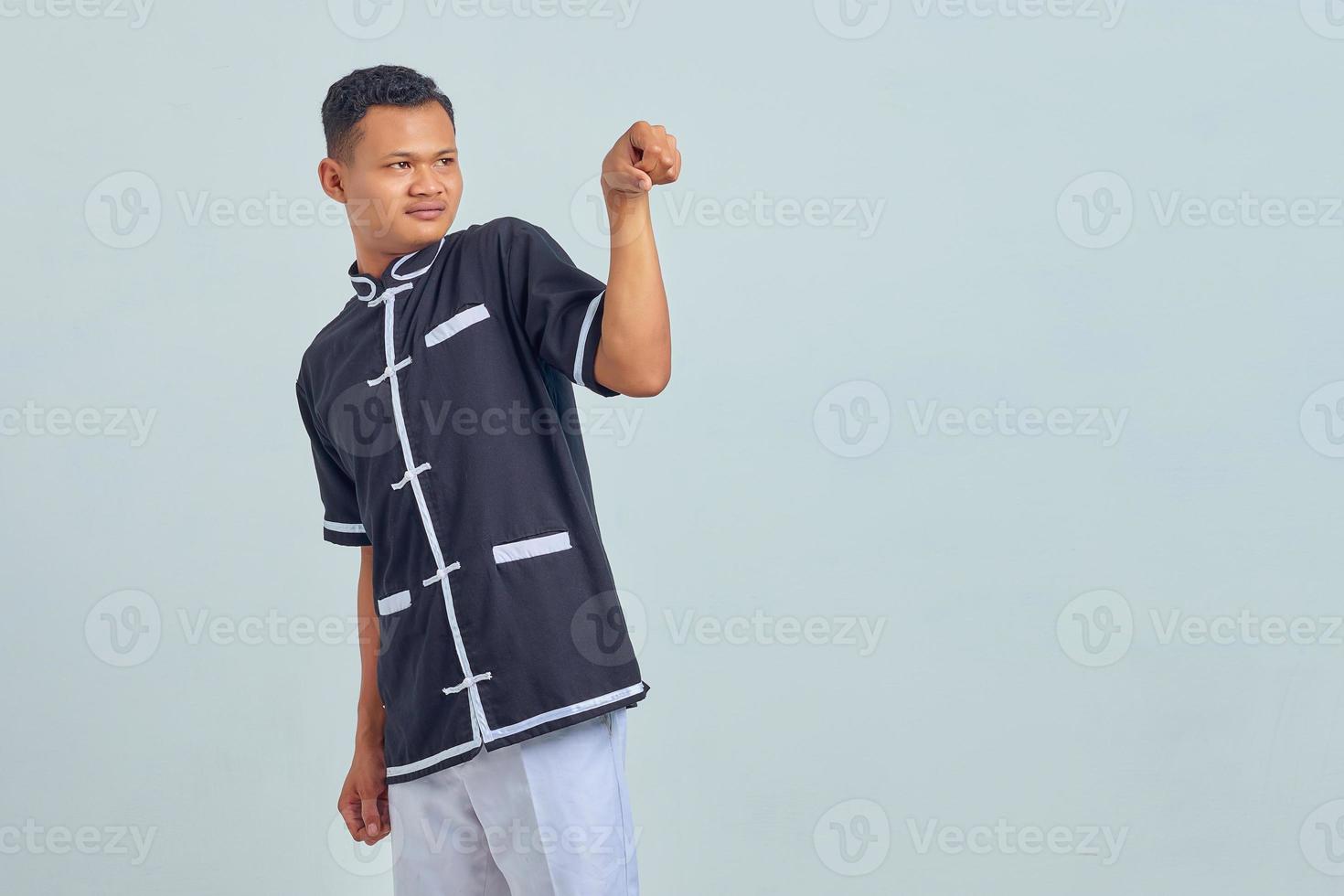 Portrait of confused asian young man wearing karate uniform showing fist gesture to empty space okay on gray background photo