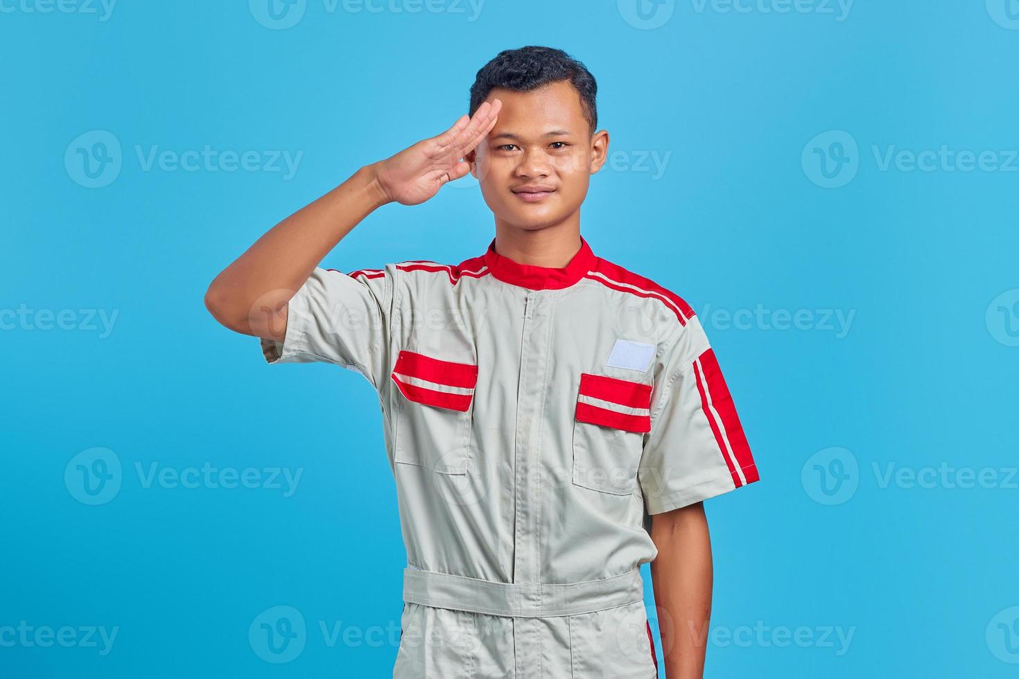Portrait of smiling young Asian mechanic making salute sign with palm isolated on blue background photo