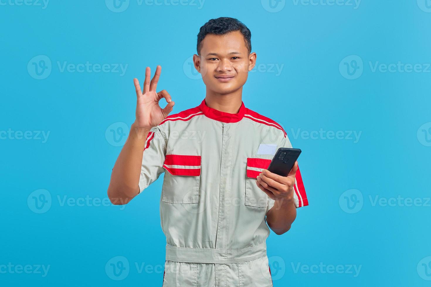 Portrait of smiling young mechanic making okay gesture and showing approval symbol isolated on blue background photo