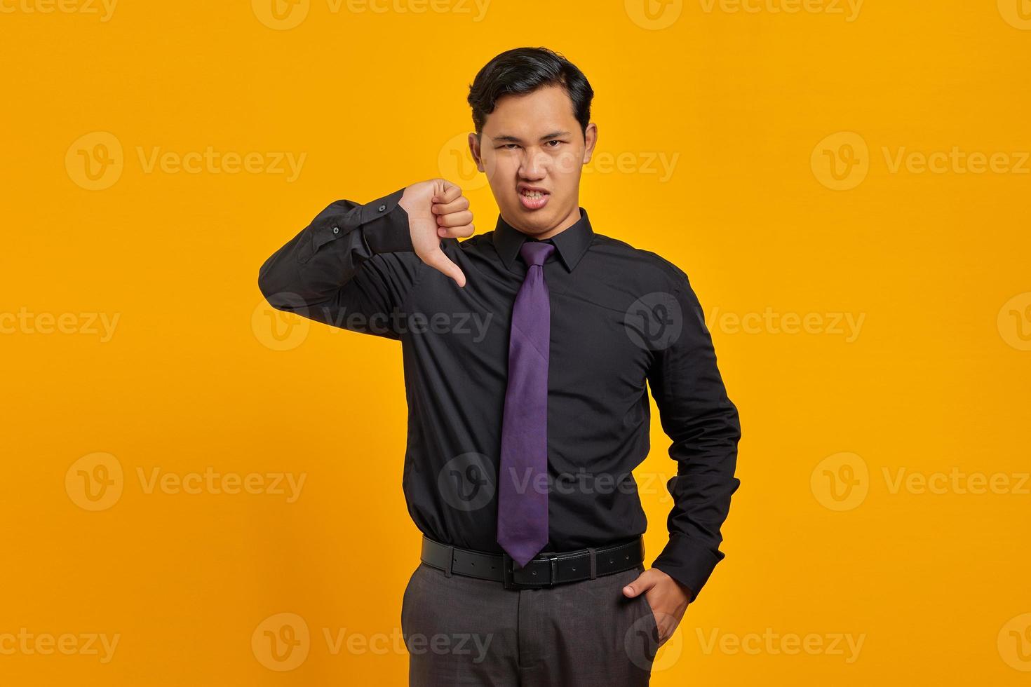 Angry young Asian businessman showing thumbs down gesture isolated on yellow background photo