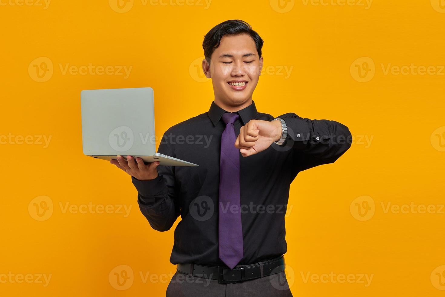 Happy young Asian businessman looking at wristwatch and holding laptop isolated on yellow background photo