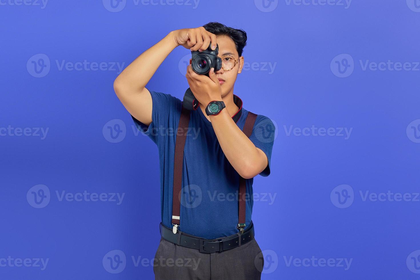 Portrait of handsome Asian young man holding professional camera and ready to take photo on purple background