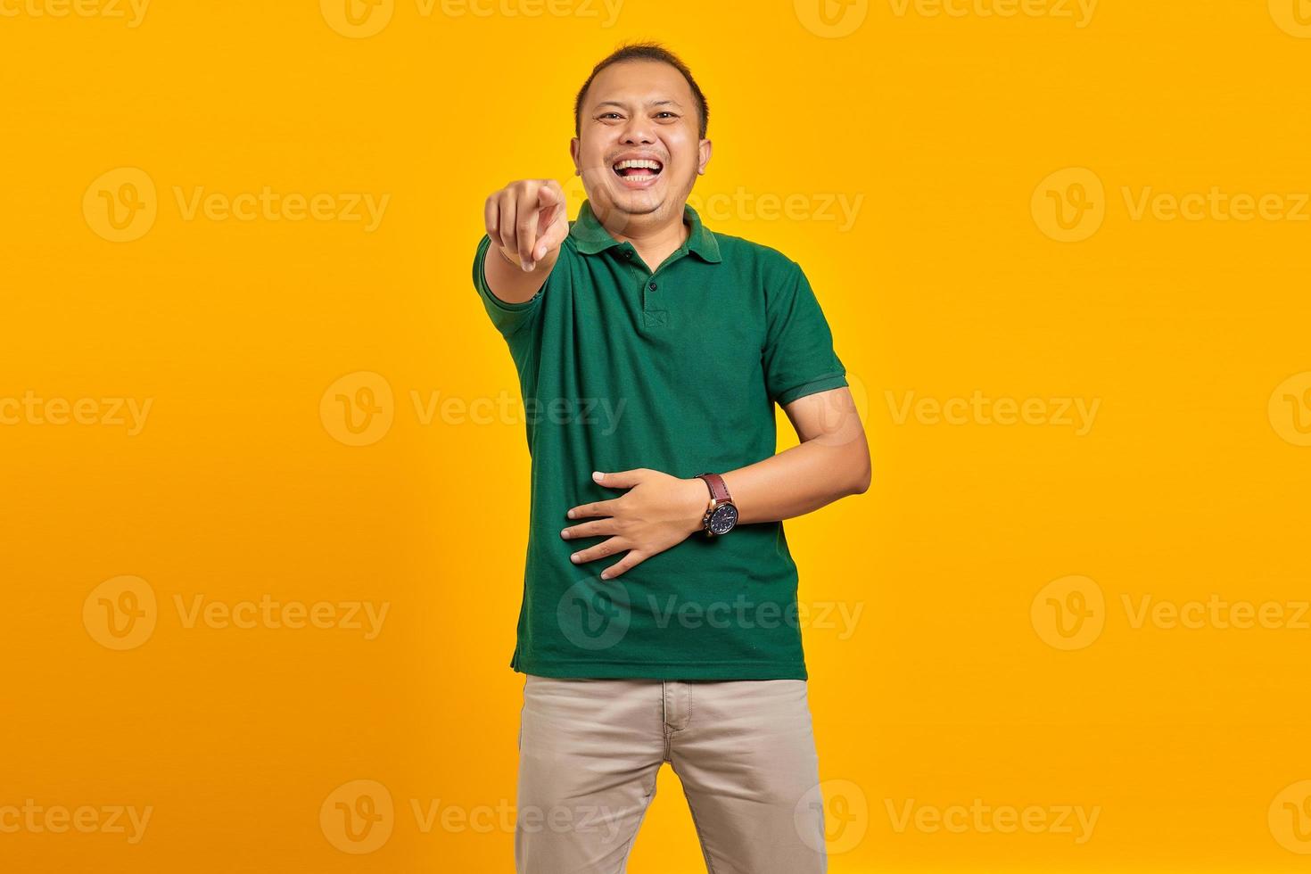 Portrait of young Asian man showing loud laughter with hand on stomach and pointing finger at camera yellow background photo