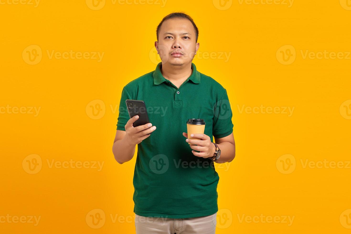 Smilling young handsome man holding smartphone and cup of coffee on yellow background photo