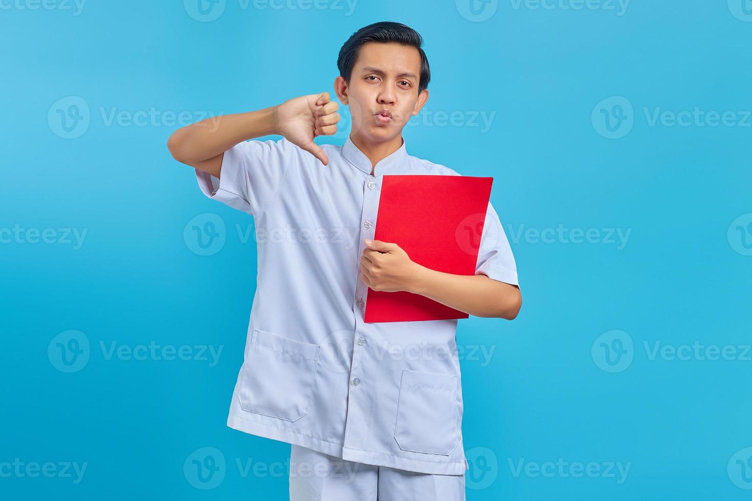 Angry young male nurse holding red folder and thumbs down gesture on blue background photo