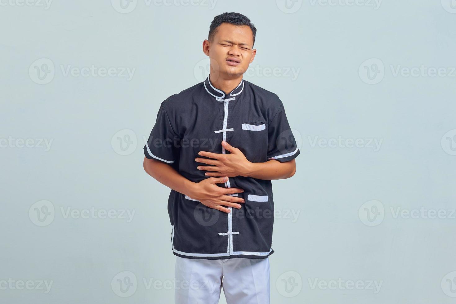 Retrato de joven asiático vistiendo uniforme de karate en el dolor con las manos en el estómago sobre fondo gris foto