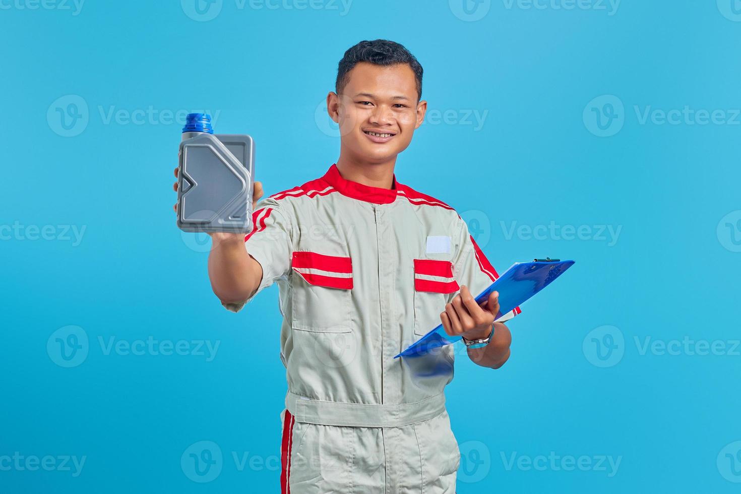 Portrait of cheerful young asian mechanic holding clipboard and showing plastic bottle of engine oil in hand over blue background photo