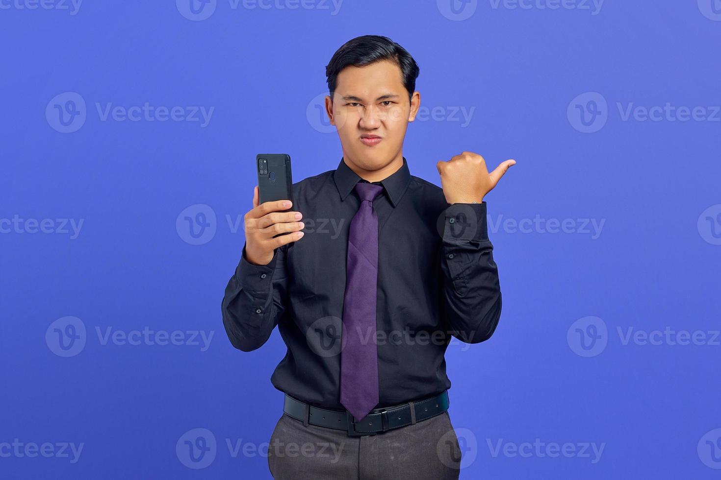 Sorprendido apuesto joven empresario apuntando a la sala de copias y sosteniendo el teléfono inteligente sobre fondo púrpura foto