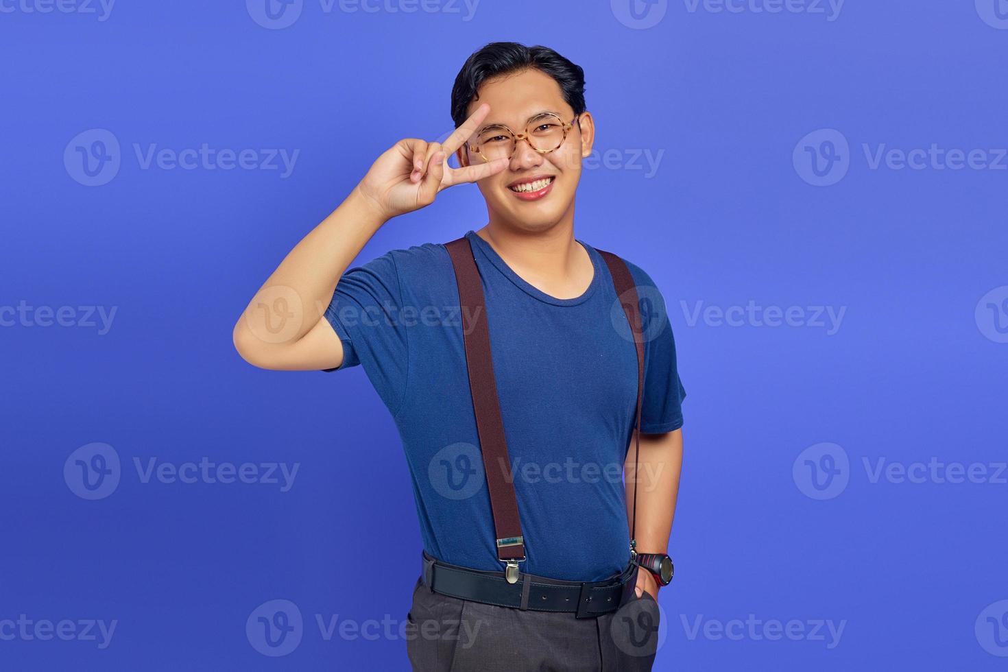 Portrait of cheerful asian man showing peace sign over eyes over purple background photo