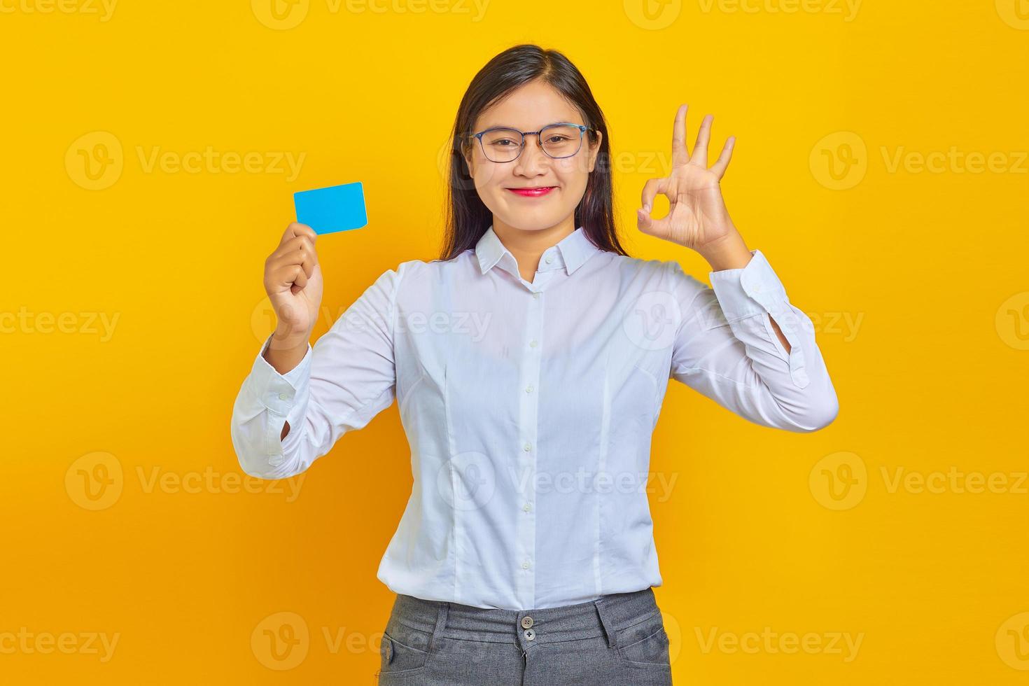 Photo of smiling Asian woman holding credit card and showing ok sign over yellow background
