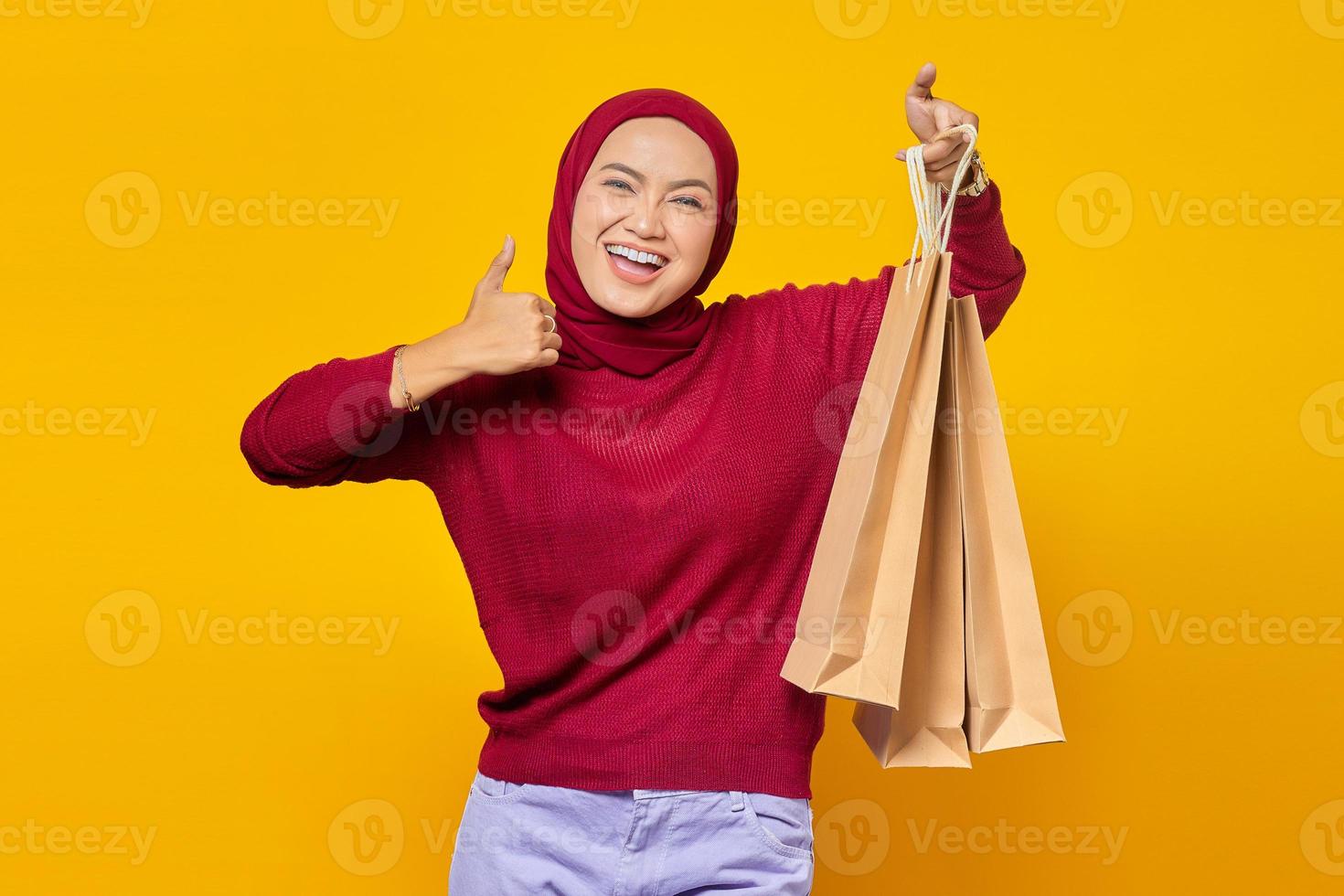 Hermosa mujer asiática mostrando algunas bolsas de la compra y dando pulgar hacia arriba con cara sonriente sobre fondo amarillo foto