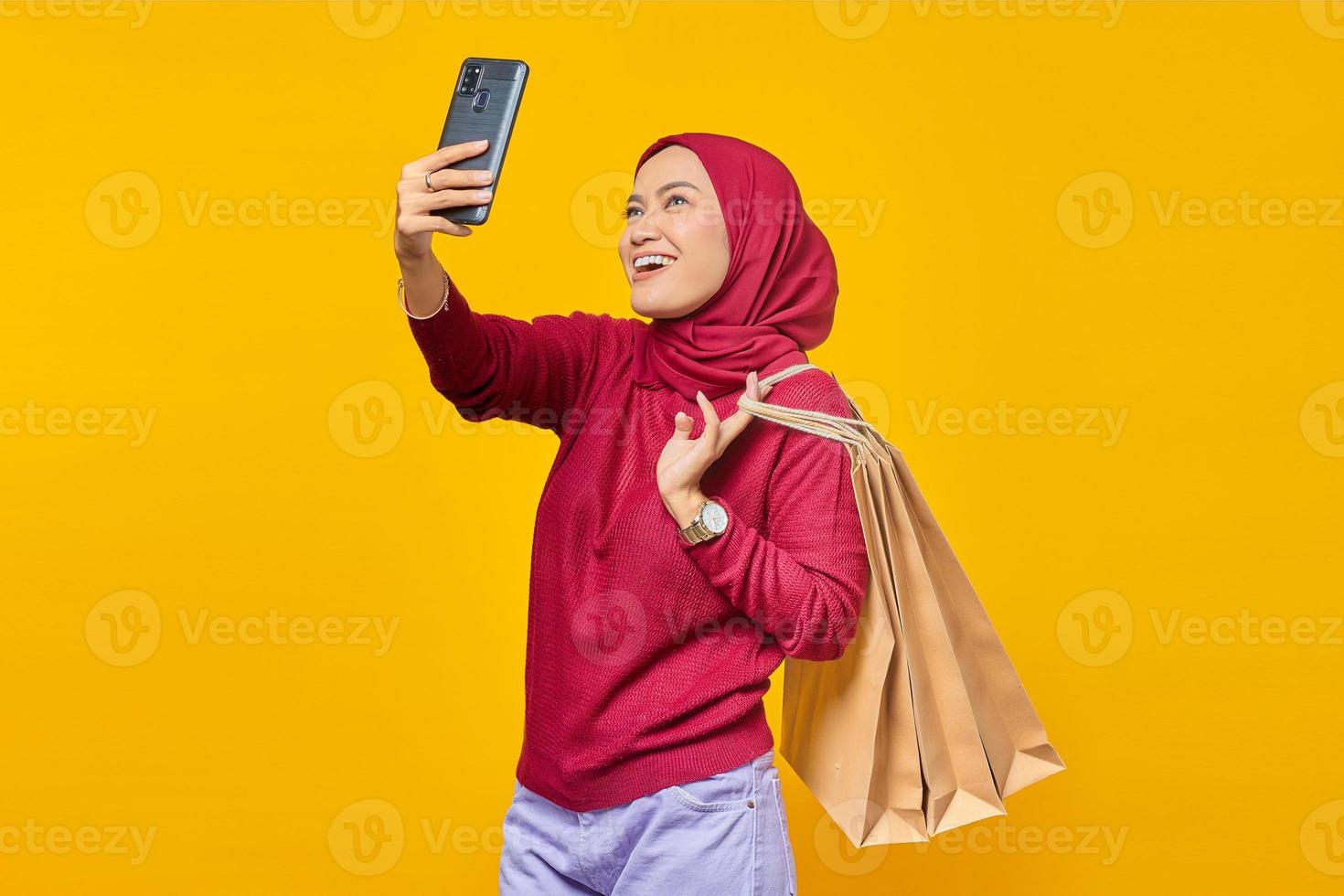 Happy young Asian woman holding shopping bags and take a selfie on mobile phone on yellow background photo