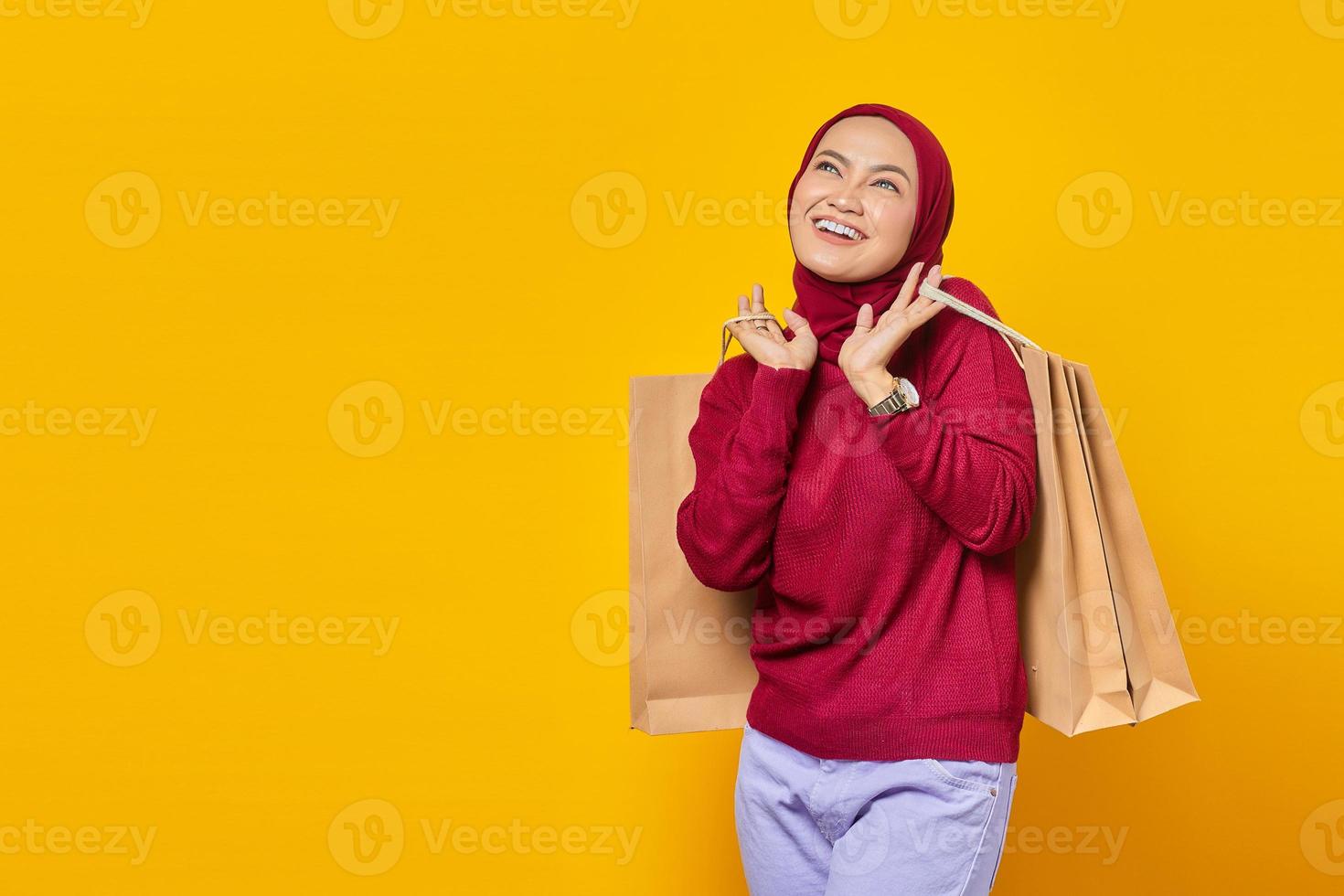 Happy young Asian woman with hands holding shopping bags and looking up on yellow background photo