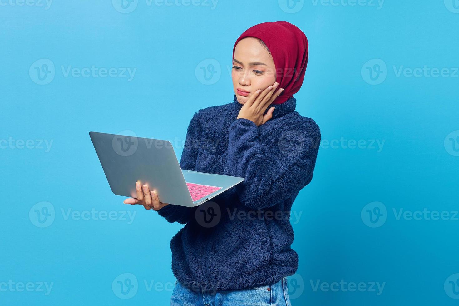 Beautiful Asian woman using laptop with confused expression looking at and holding cheek with palm photo