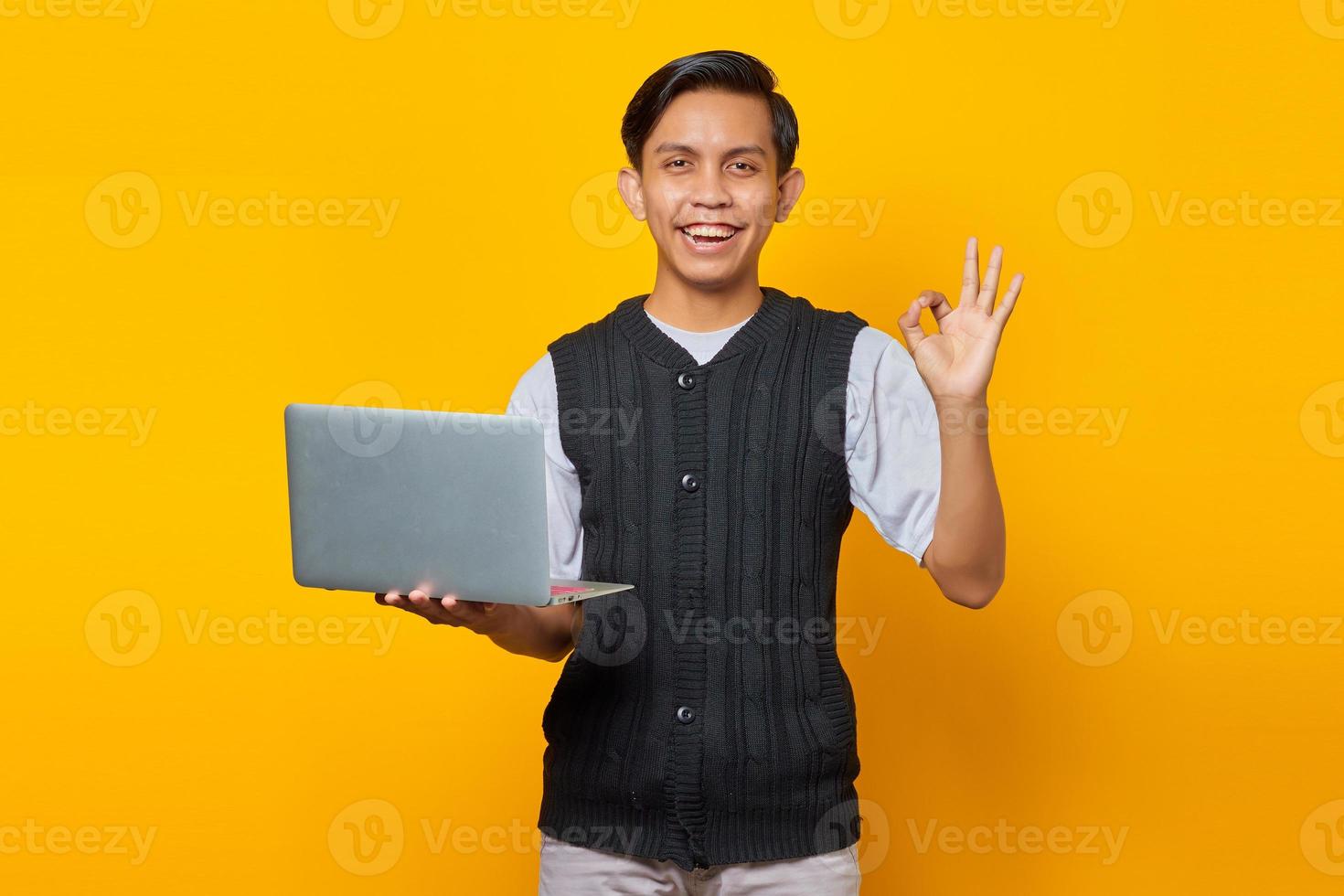 Alegre apuesto hombre asiático sosteniendo un portátil y mostrando un gesto de ok sobre fondo amarillo foto