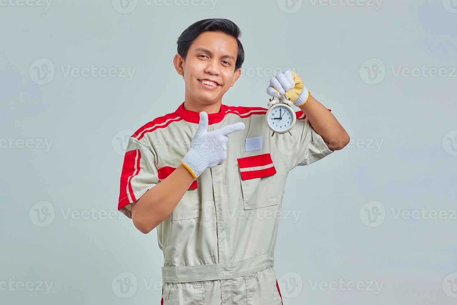 Portrait of smiling handsome man wearing mechanical uniform holding alarm clock on gray background photo