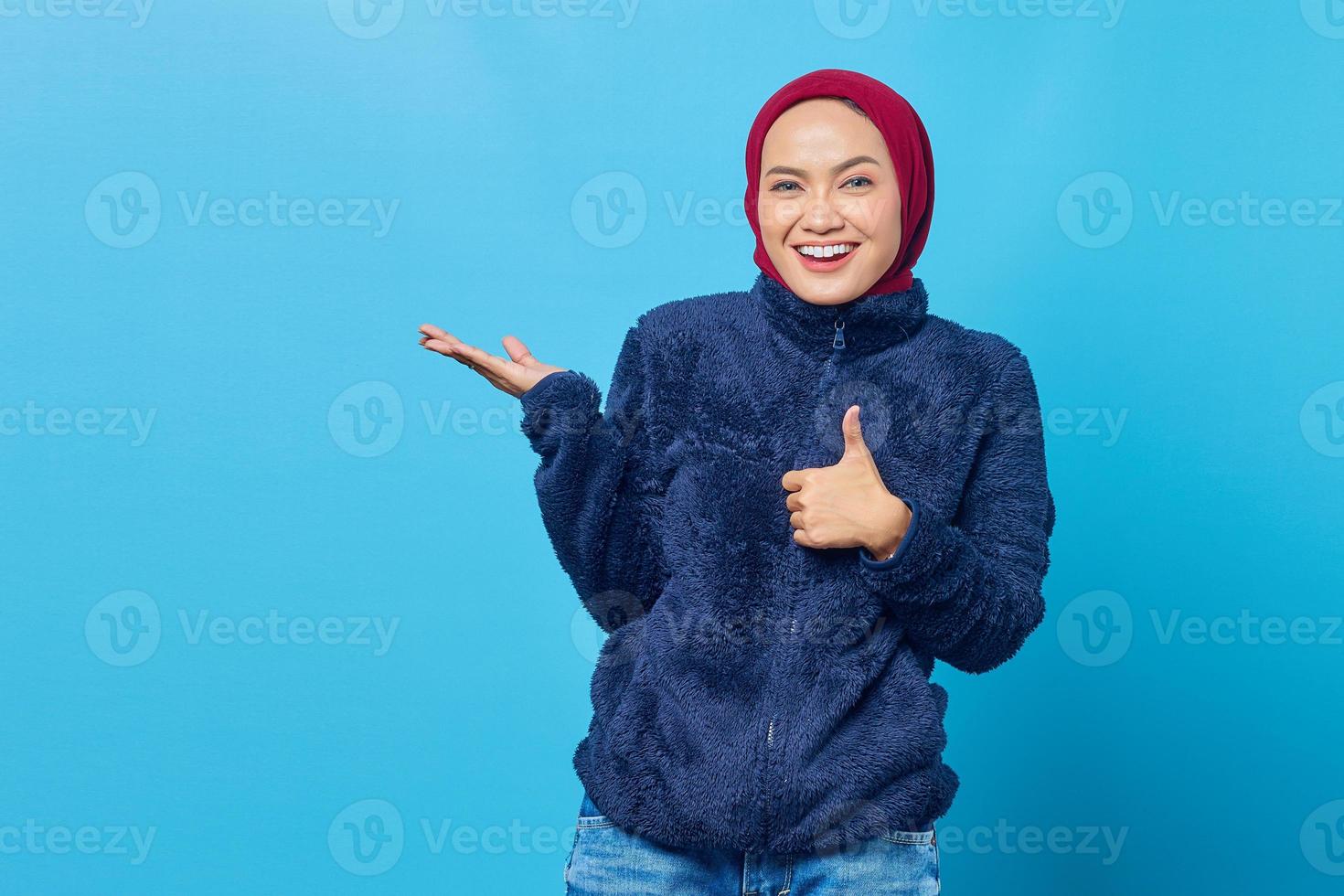 Portrait of smiling young Asian woman giving thumbs up and pointing to empty space with palms on blue background photo