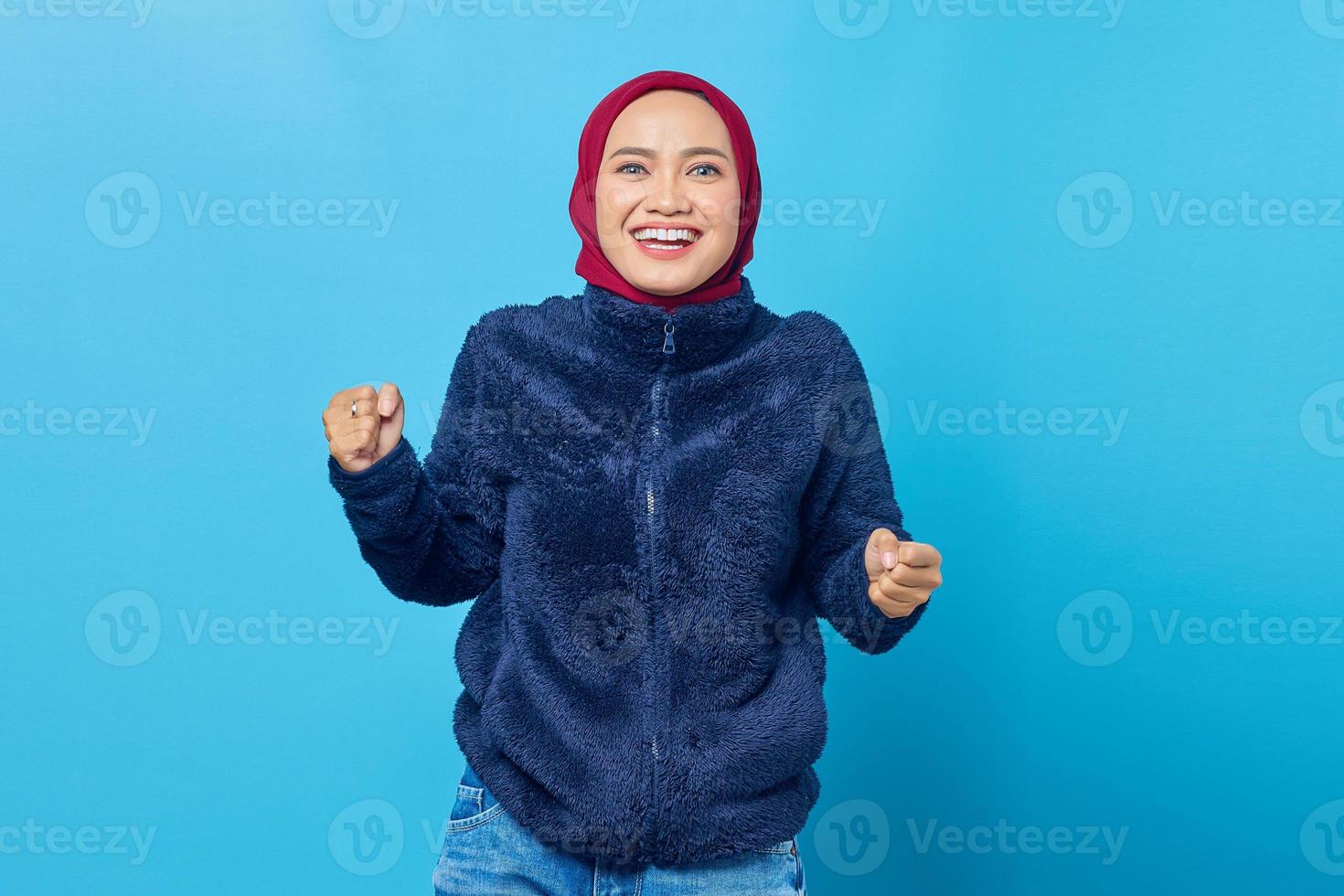 Emocionada mujer asiática celebrando el éxito con dos puños en el aire sobre fondo azul. foto