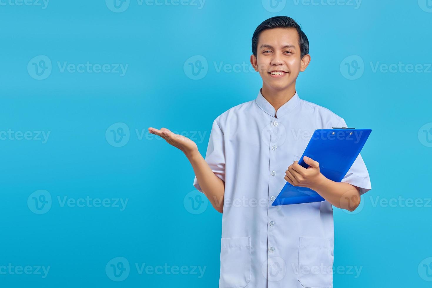 Retrato de alegre enfermero sosteniendo el portapapeles y apuntando hacia los lados con las palmas sobre fondo azul. foto