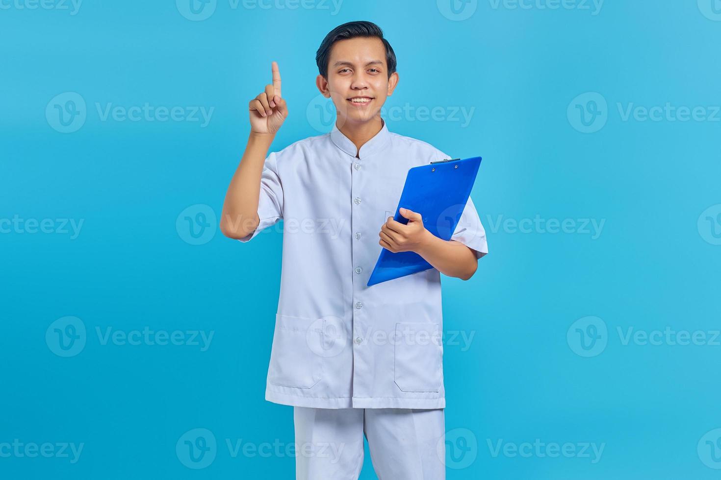 Portrait of cheerful male nurse holding clipboard standing and pointing up on blue background photo