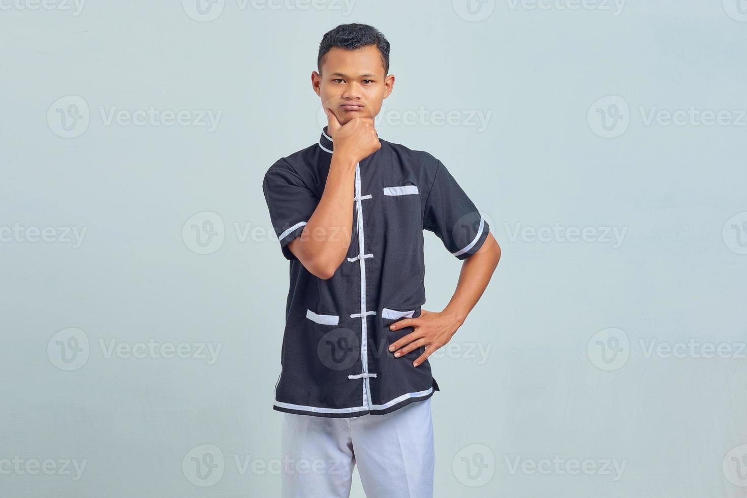 Retrato de pensativo joven asiático vistiendo uniforme de karate frotando la barbilla y pensando en pregunta aislada sobre fondo gris foto
