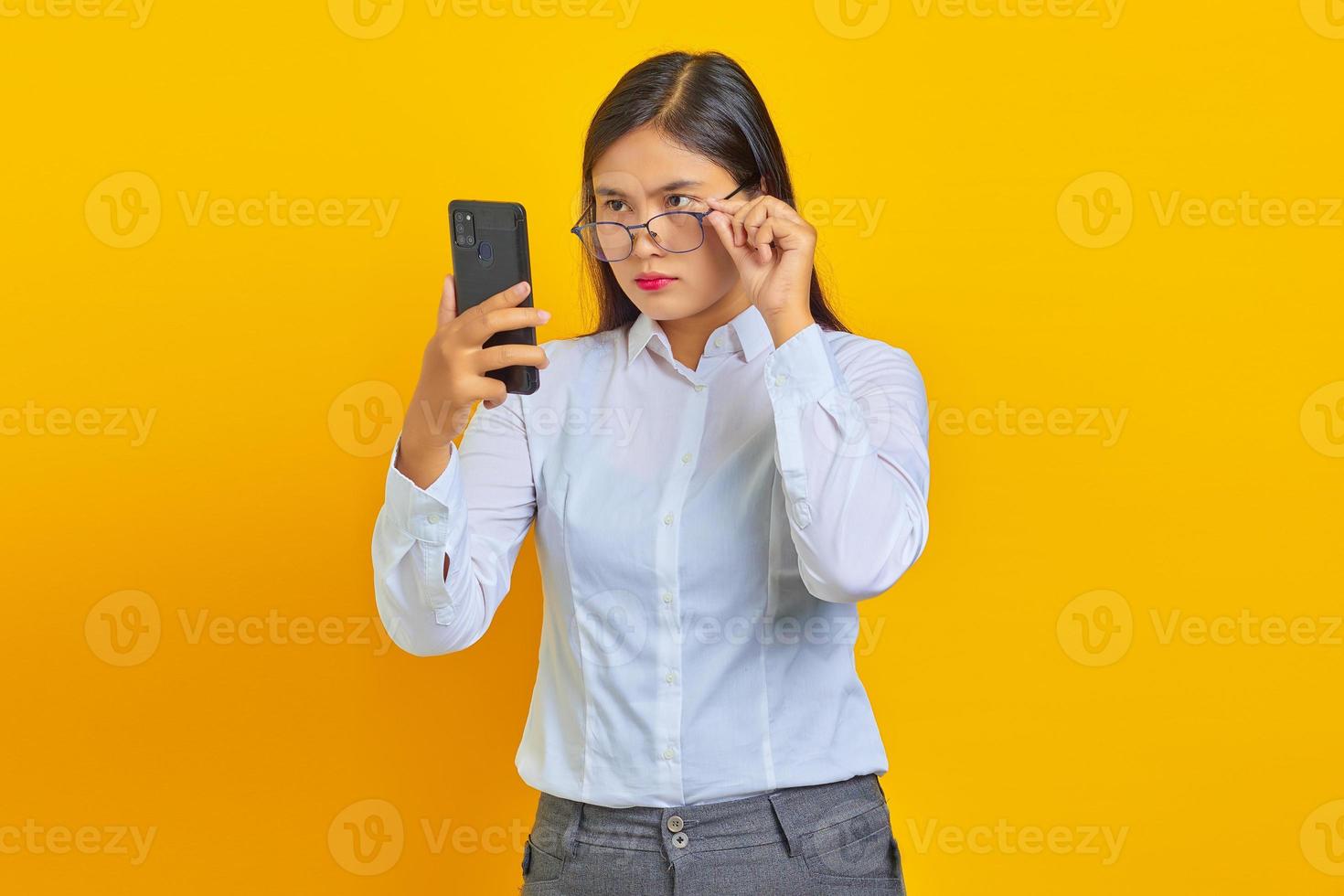 Mujer asiática joven enojada que usa el teléfono móvil y que toma las gafas aisladas sobre fondo amarillo foto