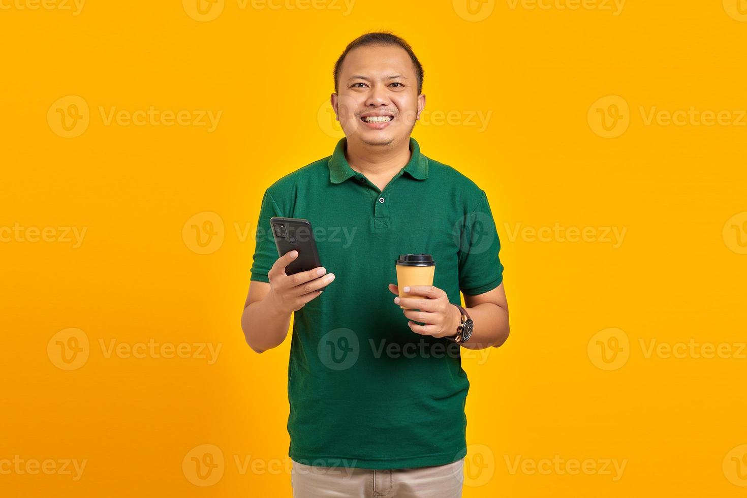 Cheerful young handsome man holding smartphone and cup of coffee on yellow background photo