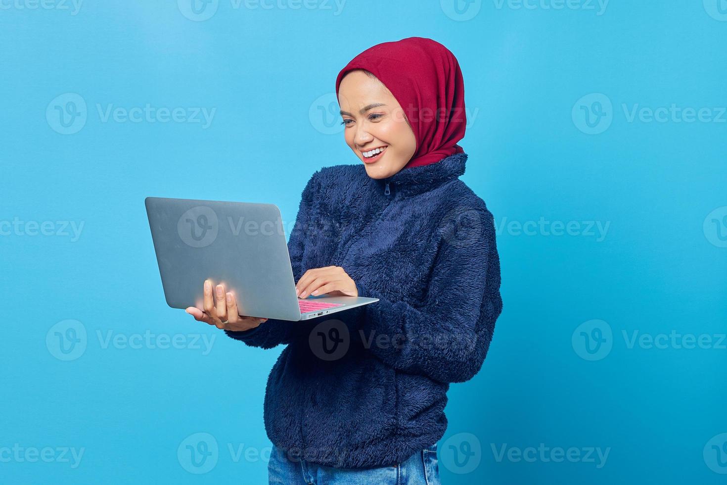 Foto de mujer asiática alegre usando laptop para compras en línea sobre fondo azul.