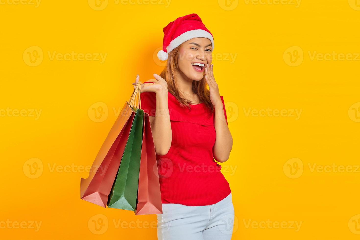 Emocionada joven asiática en santa claus hat sosteniendo bolsas de la compra y mirando a la cámara aislada sobre fondo amarillo foto