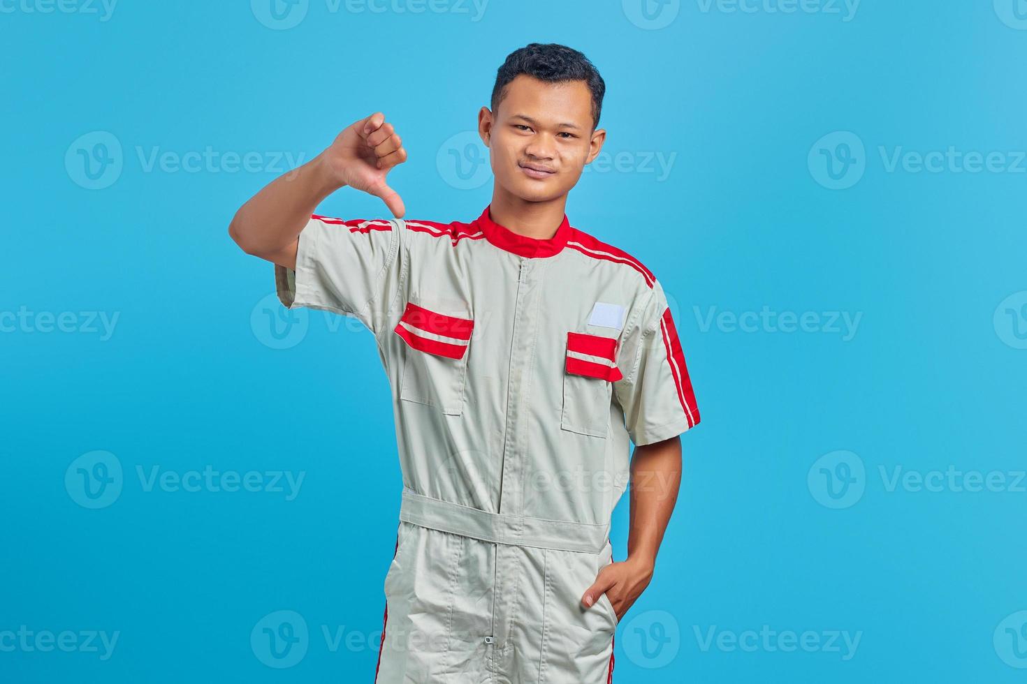 Angry young mechanic showing refusal with thumbs down gesture isolated on blue background photo